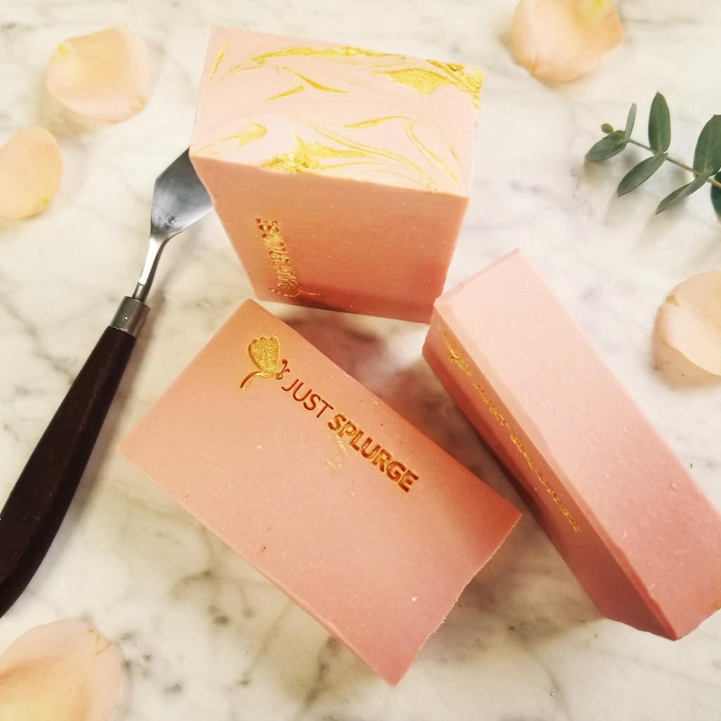 Three peach-pink soap bars with gold Just Splurge details on warm ivory marble, framed by peach petals and greenery.