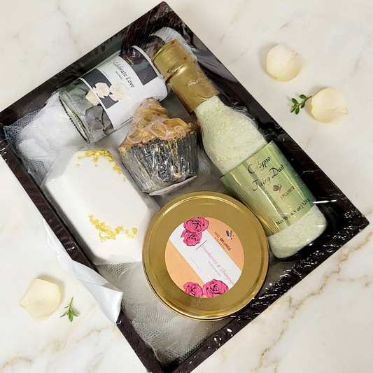 Black gift tray with wrapped bath and body products and a gold tin on ivory marble with cream petals and green accents.