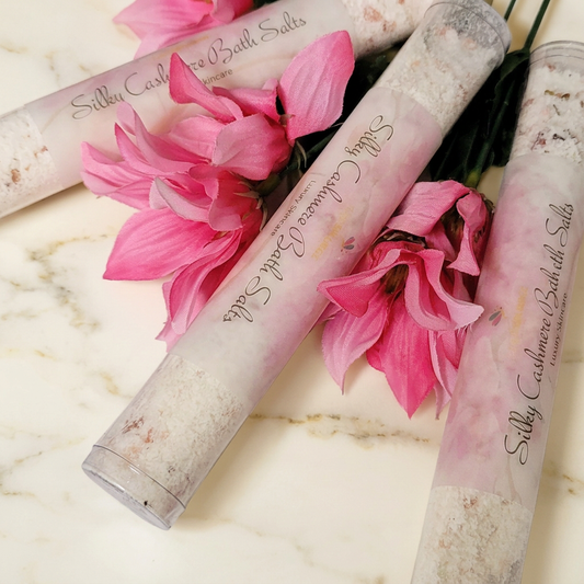 Clear tubes of Silky Cashmere Bath Salts with pink labels and pink flowers on warm ivory marble.