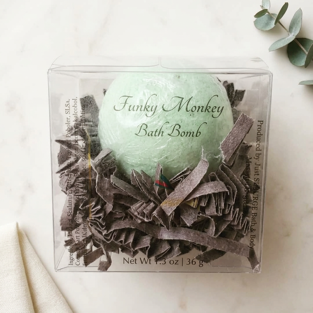 Pale green Funky Monkey bath bomb in a clear gift box with brown paper filling, on ivory marble with linen and eucalyptus.