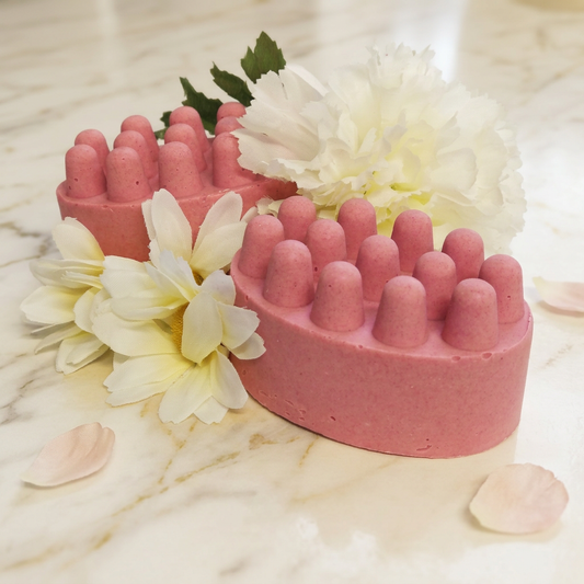 Two pink oval Strawberry Massage Bars with rounded knobs, cream flowers and blush petals on warm ivory marble.