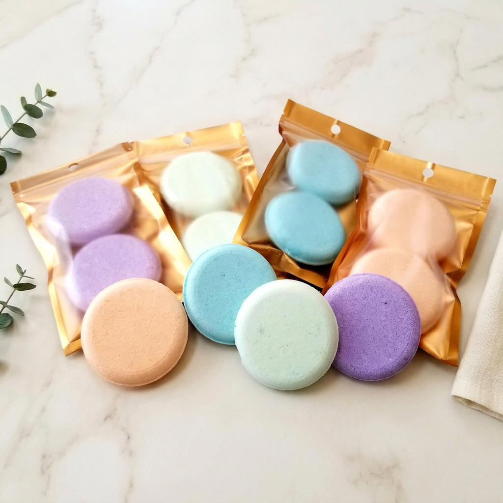 Pastel shower steamer tablets and gold pouches on ivory marble with eucalyptus and ivory linen.