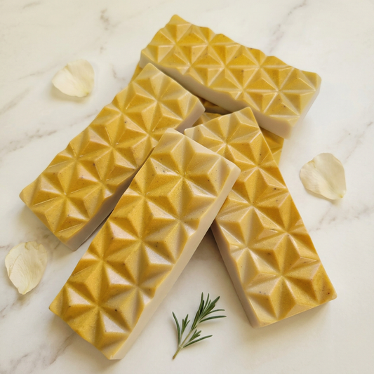 Gold-toned geometric wax melt bars with ivory sides arranged on warm marble with cream petals and a green sprig.