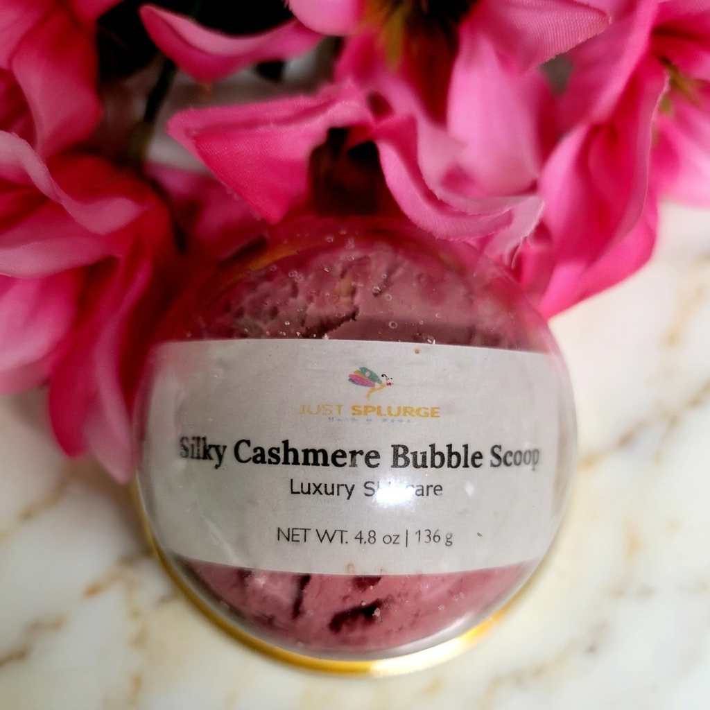 Clear round container of pink Silky Cashmere Bubble Scoop with pink flowers on warm ivory marble.