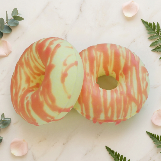 Two cream donut-shaped Calypso bath bombs with coral pink stripes on warm ivory marble framed by blush petals and greenery.