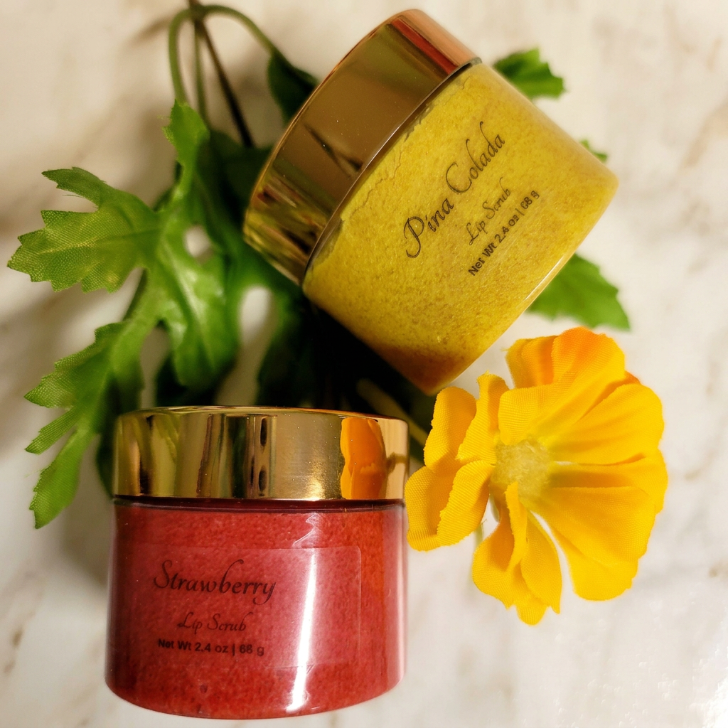 Yellow Pina Colada and red Strawberry lip scrub jars with gold lids, a yellow flower and greenery on warm ivory marble.