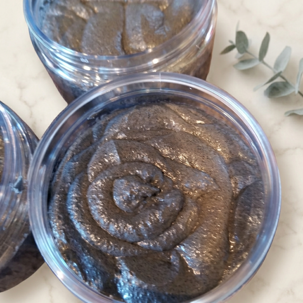 Open clear jars of dark gray-brown swirled sugar polish on warm ivory marble with a eucalyptus accent.